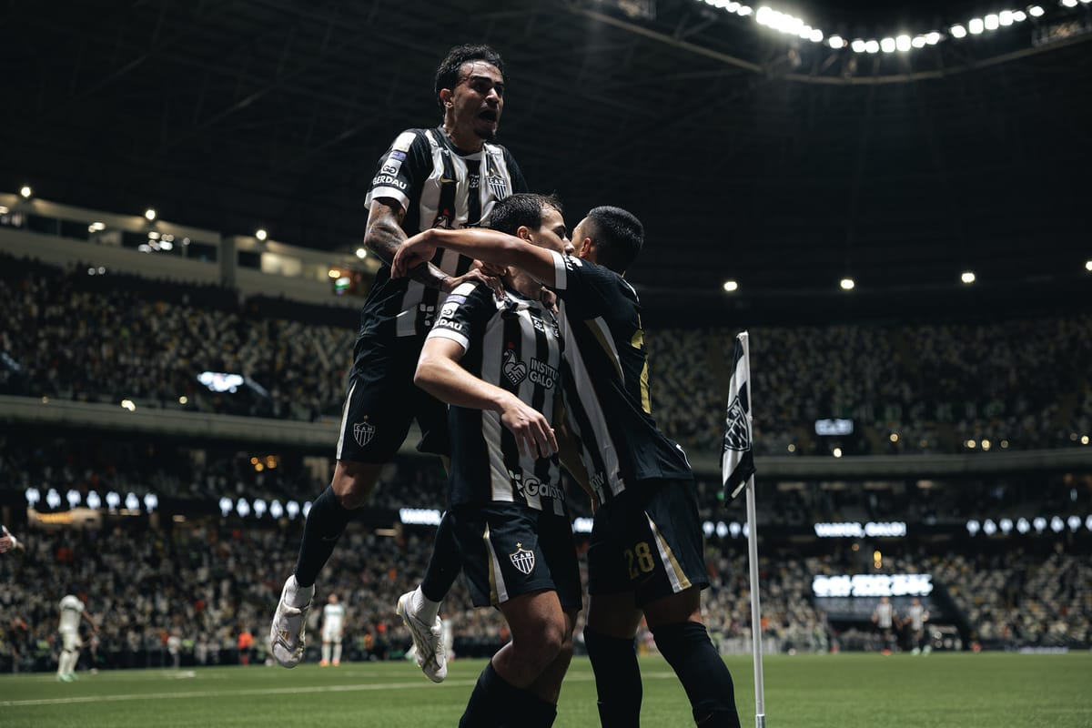 Jogadores do Atlético-MG, de camisa listrada preta e branca, se abraçam e um deles salta sobre os outros ao comemorar gol, perto da bandeira de escanteio, com a Arena MRV lotada ao fundo