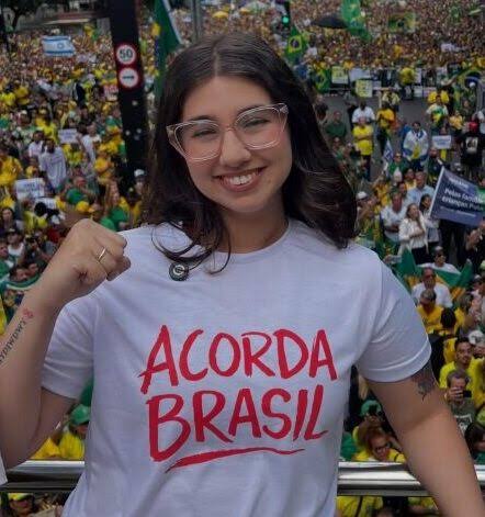 Eduarda Campopiano, de óculos e camiseta branca com a frase Acorda Brasil, sorri com o punho erguido diante de uma multidão de verde e amarelo
