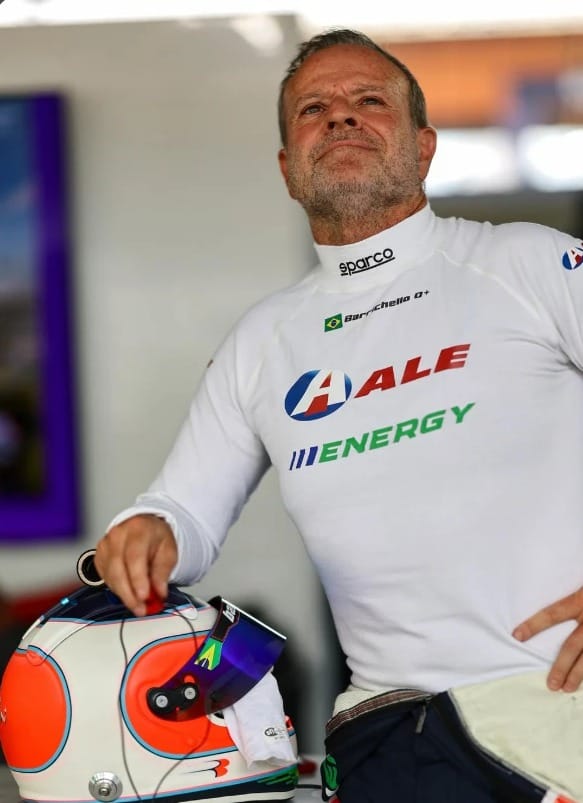 Rubens Barrichello, de macacão branco, segura o capacete e olha para cima nos boxes