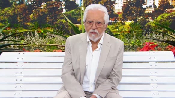 Carlos Alberto de Nóbrega, de cabelo e barba brancos, óculos e terno bege, sentado no banco do cenário de A Praça é Nossa