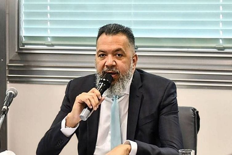 Márcio Canella, de barba grisalha e terno escuro, fala ao microfone sentado a uma mesa