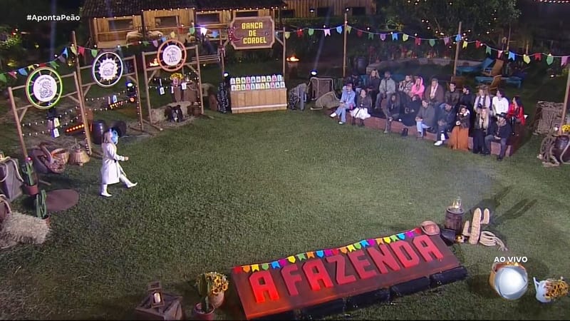 Peões de A Fazenda 18 sentados ao lado do gramado iluminado, à noite, durante a dinâmica, com o letreiro vermelho do programa