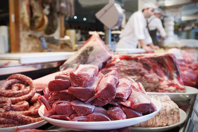 Bifes de carne vermelha empilhados num prato no balcão de um açougue, com açougueiros trabalhando ao fundo