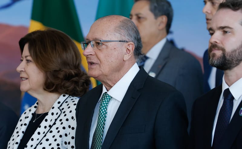 Geraldo Alckmin, de óculos, terno escuro e gravata verde, em primeiro plano, ao lado de uma mulher de blusa de bolinhas, com bandeiras do Brasil ao fundo