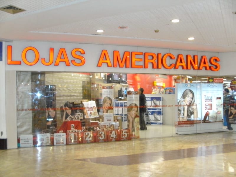 Entrada de uma loja Lojas Americanas em shopping, com o nome da rede em letras laranja sobre a vitrine
