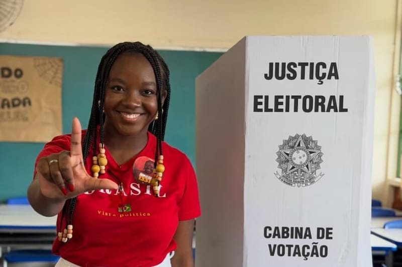 Ana Elisa, de camiseta vermelha, sorri e faz o L com a mão ao lado da cabine de votação da Justiça Eleitoral