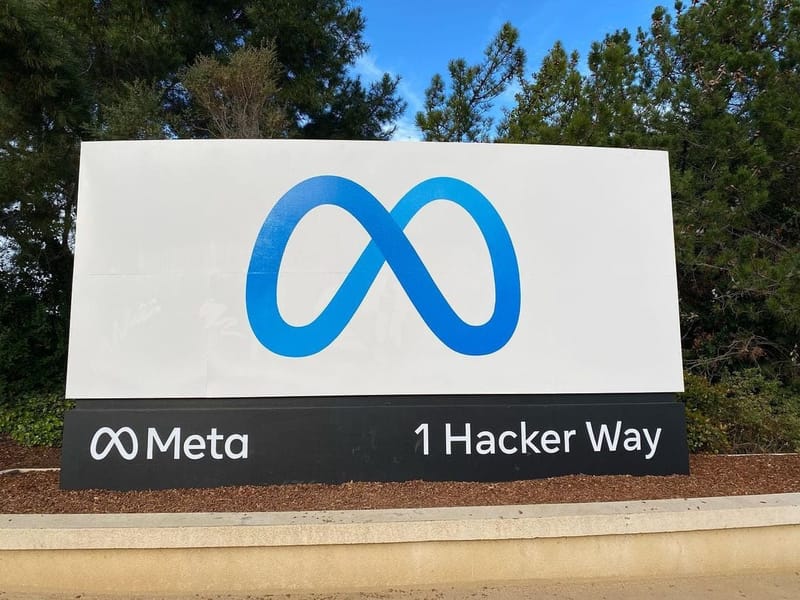 Placa branca com o símbolo azul de infinito da Meta e o endereço 1 Hacker Way, cercada por árvores