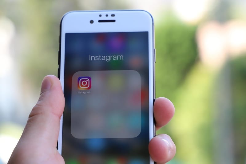 Mão segura um smartphone de moldura branca cuja tela mostra o ícone do aplicativo Instagram dentro de uma pasta
