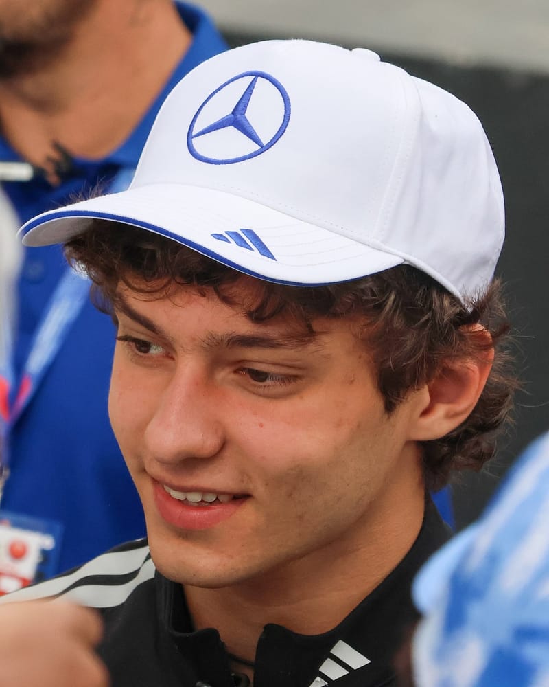 Kimi Antonelli sorri de boné branco com o símbolo da Mercedes, cercado de pessoas no paddock