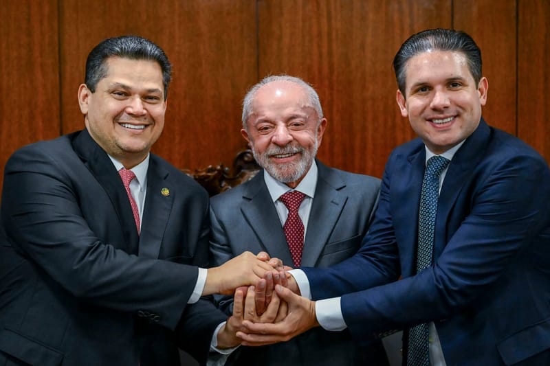 Lula, de terno cinza e gravata vermelha, sorri entre Davi Alcolumbre e Hugo Motta, que unem as mãos aos dele