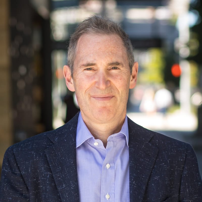 Andy Jassy, de cabelo curto grisalho, paletó azul-escuro e camisa azul-clara, sorri de leve em retrato de frente, com a rua desfocada ao fundo