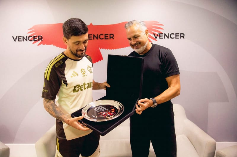 O meia Arrascaeta, de camisa do Flamengo, segura uma bandeja de homenagem ao lado de um homem de barba grisalha