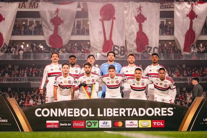 Jogadores do Flamengo, de uniforme branco, posam enfileirados em campo diante de um painel da Libertadores e de faixas da torcida nas arquibancadas