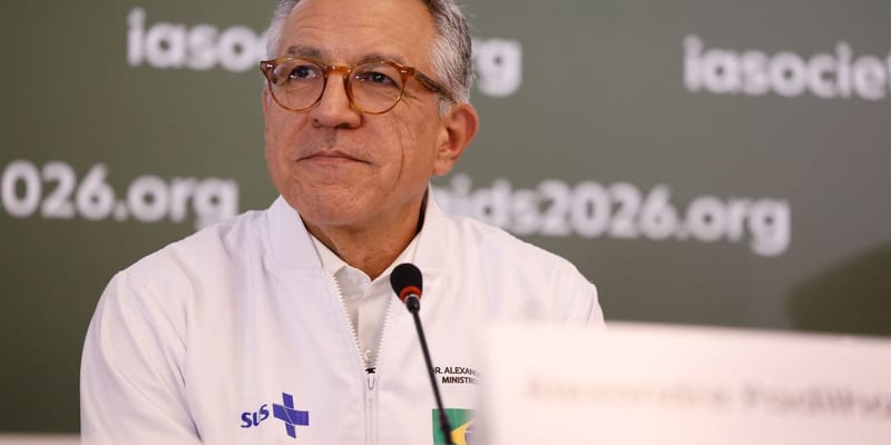 Alexandre Padilha, de óculos de armação marrom e jaqueta branca do SUS, fala diante de um microfone, com painéis da conferência ao fundo