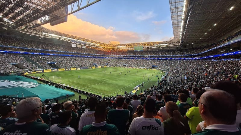 Arquibancadas do estádio do Palmeiras cheias de torcedores sob a cobertura, com o gramado ao centro e céu ao entardecer