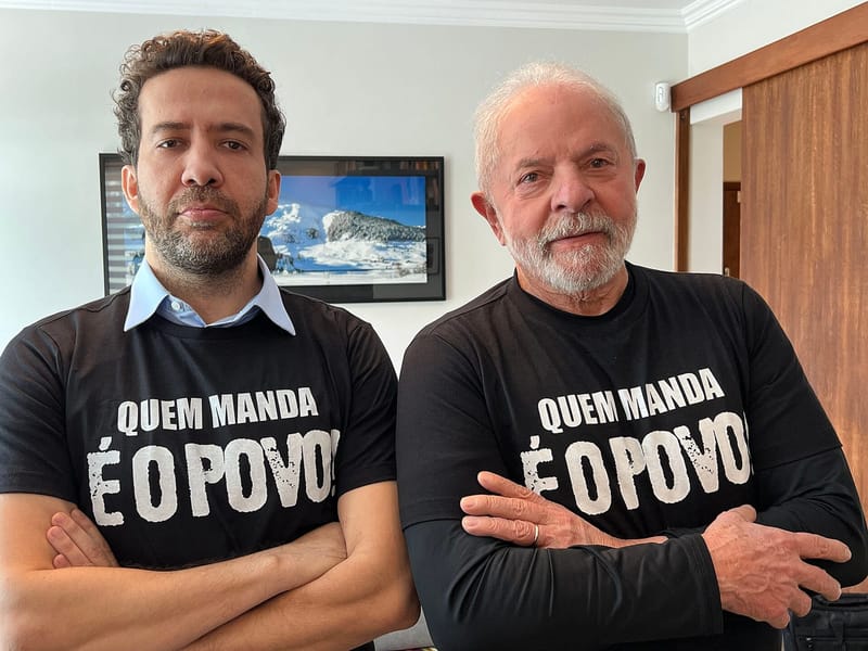 Lula e André Janones lado a lado, de braços cruzados, com camisetas pretas escritas Quem manda é o povo
