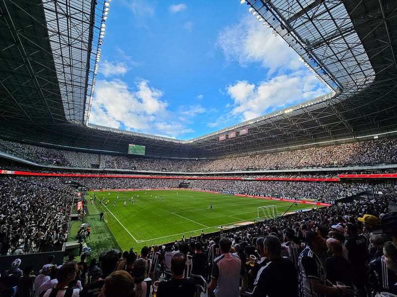 Interior da Arena MRV com a cobertura aberta ao céu azul, a torcida nas arquibancadas e o jogo em andamento no gramado