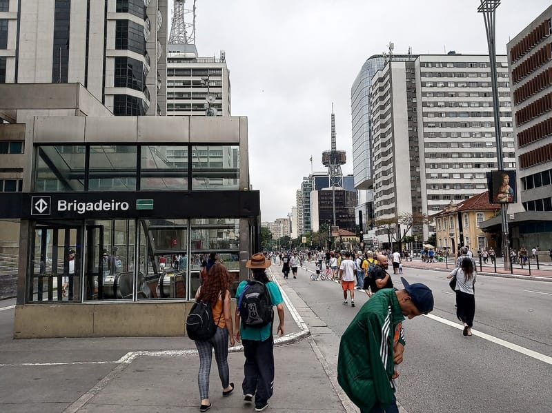 Entrada da estação Brigadeiro do metrô na Avenida Paulista, com pedestres na calçada e edifícios ao fundo