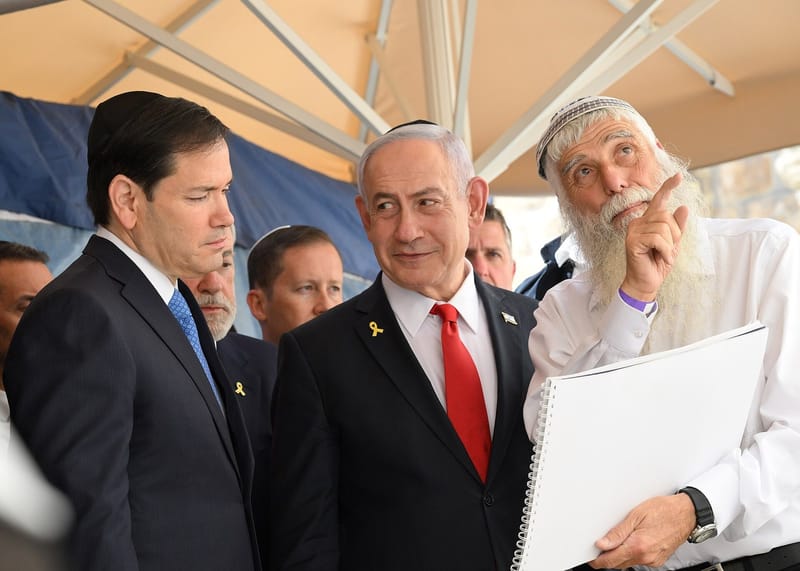 Benjamin Netanyahu, de quipá, terno escuro e gravata vermelha, olha de lado ao lado de outros homens durante uma visita oficial