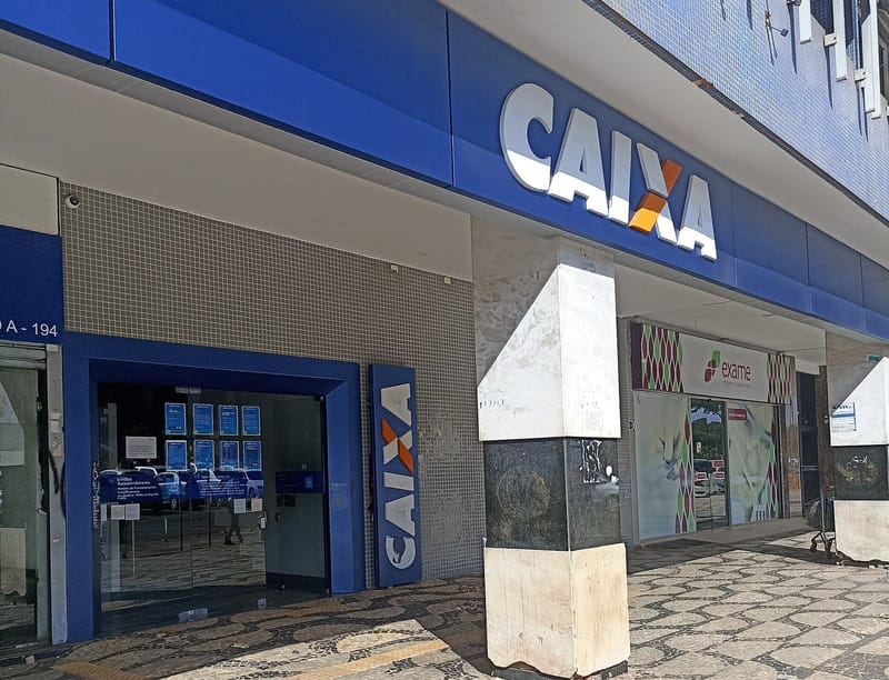 Fachada de agência da Caixa Econômica Federal, com o logotipo azul e laranja sobre a entrada e calçada de pedras portuguesas