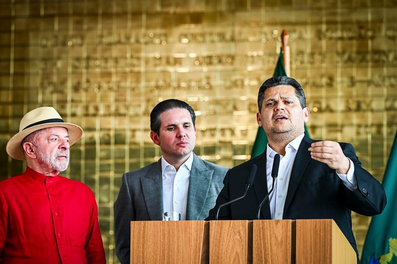 Davi Alcolumbre, de terno escuro e camisa clara, fala diante de dois microfones ao lado de Hugo Motta e de Lula, que veste camisa vermelha e chapéu de palha