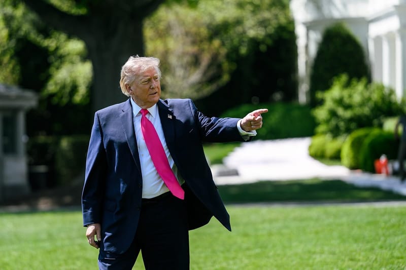 Donald Trump, de terno azul-marinho e gravata rosa, aponta com o braço esticado no gramado da Casa Branca