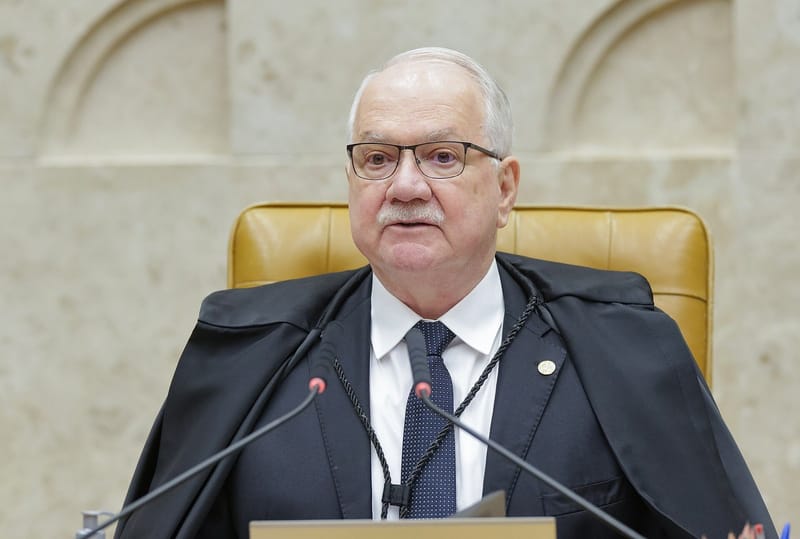 Edson Fachin, de óculos, toga preta e gravata azul-marinho, ao microfone, na cadeira central do plenário, sob o brasão da República