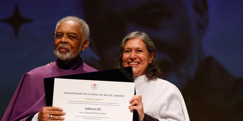 Gilberto Gil, de barba branca e beca roxa, sorri segurando o diploma da Uerj ao lado da reitora, de beca branca