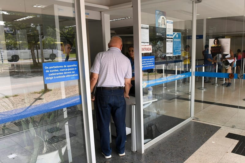 Porta de vidro da agência do INSS, com sinalização azul de entrada e um homem de camisa clara entrando