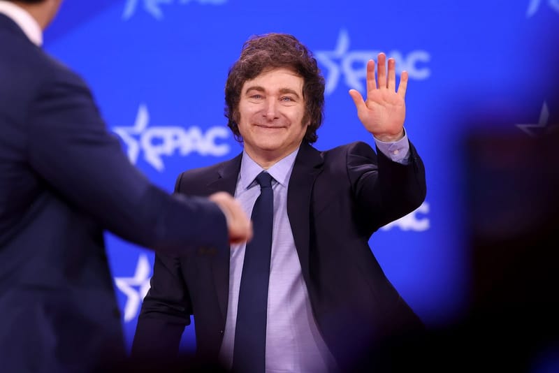 Javier Milei, de terno escuro e gravata, acena com a mão direita diante de painel azul