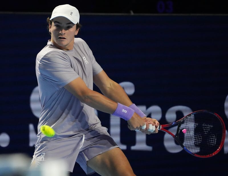 João Fonseca, de boné e camiseta cinza e munhequeiras roxas, rebate uma bola amarela de backhand em quadra azul-escura