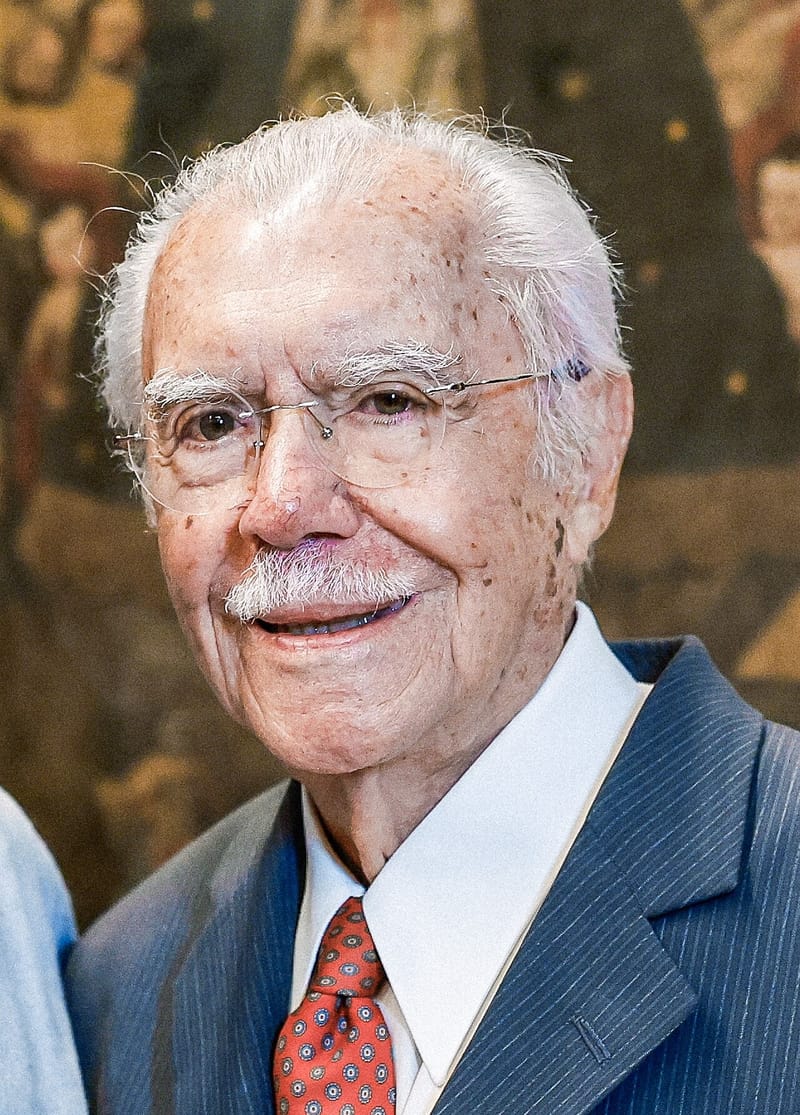 Retrato de José Sarney, de óculos sem aro, cabelos brancos e sorriso discreto, com terno azul listrado e gravata vermelha