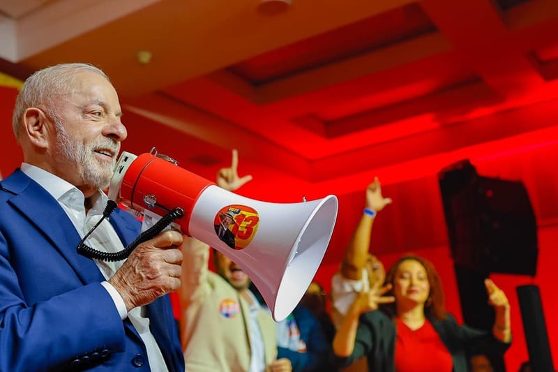 Lula, de blazer azul e camisa branca, fala com um megafone vermelho e branco em um salão iluminado de vermelho, com apoiadores ao fundo