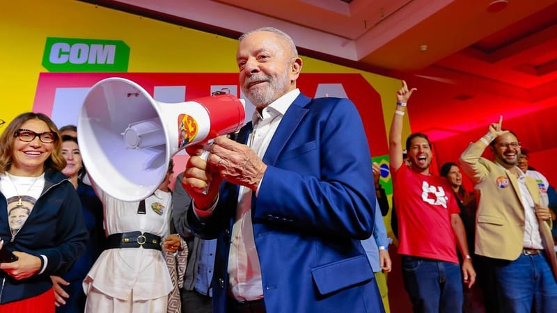 Lula, de blazer azul, segura um megafone e sorri diante de um painel amarelo e vermelho, cercado de apoiadores