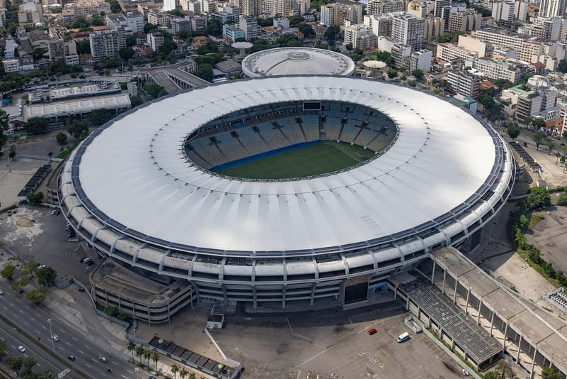 Vista de cima do Maracanã, com a cobertura branca circular, o gramado verde ao centro e prédios da zona norte do Rio ao redor