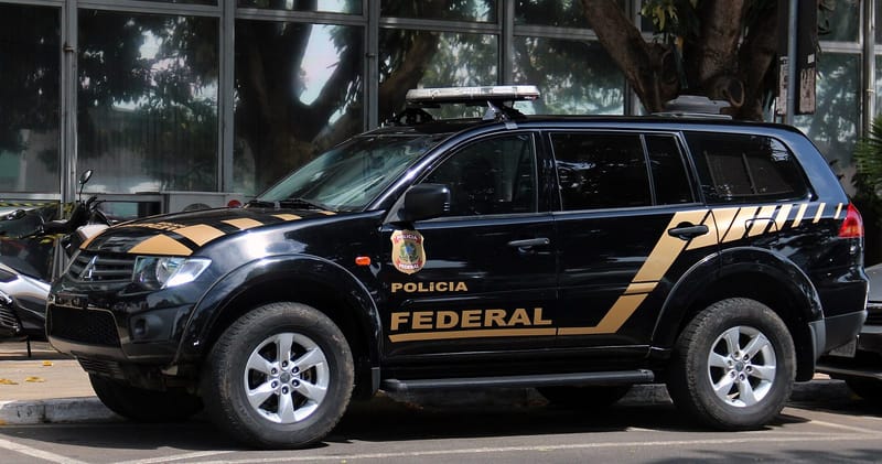 Caminhonete preta da Polícia Federal, com faixas douradas e o brasão na porta, estacionada diante de um prédio