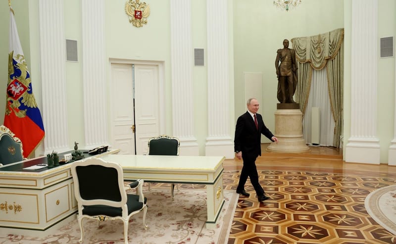 Vladimir Putin, de terno escuro, atravessa a pé um salão do Kremlin de piso de madeira, com a bandeira russa ao fundo