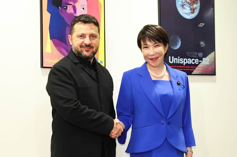 Volodymyr Zelensky, de roupa preta, aperta a mão de uma mulher de terninho azul-royal diante de dois cartazes coloridos