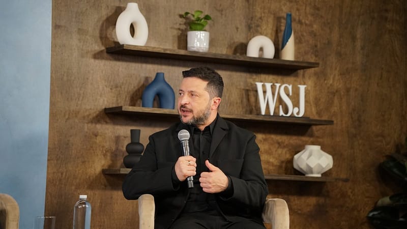 Volodymyr Zelensky, de preto, fala ao microfone e gesticula com a mão direita diante de uma parede de madeira com prateleiras