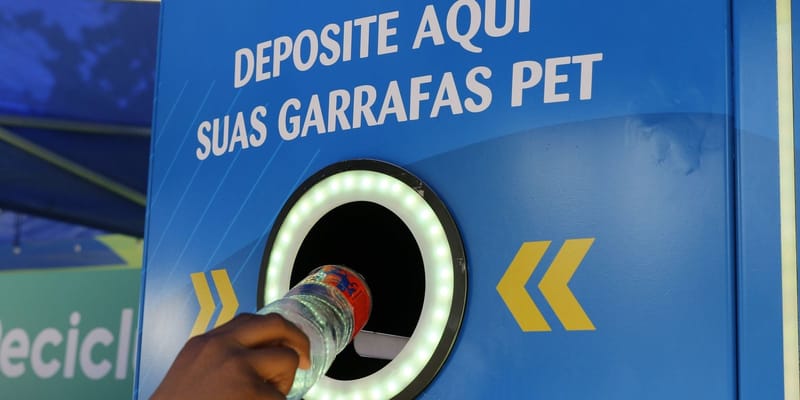 Mão insere uma garrafa PET em máquina de coleta azul com o aviso "Deposite aqui suas garrafas PET" e um anel de luz verde ao redor da abertura