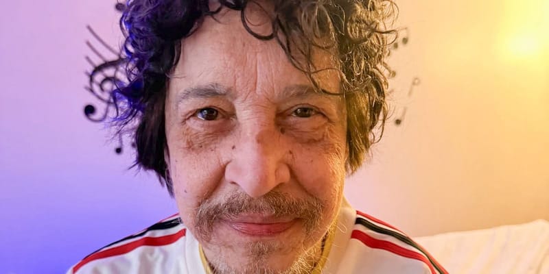 Benito Di Paula, de cabelos cacheados e bigode grisalho, sorri para a câmera usando uma camisa branca com listras vermelhas e pretas