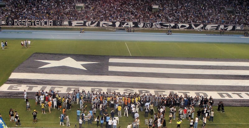 Bandeira gigante do Botafogo estendida sobre o gramado do Estádio Nilton Santos, com torcedores e jogadores ao redor