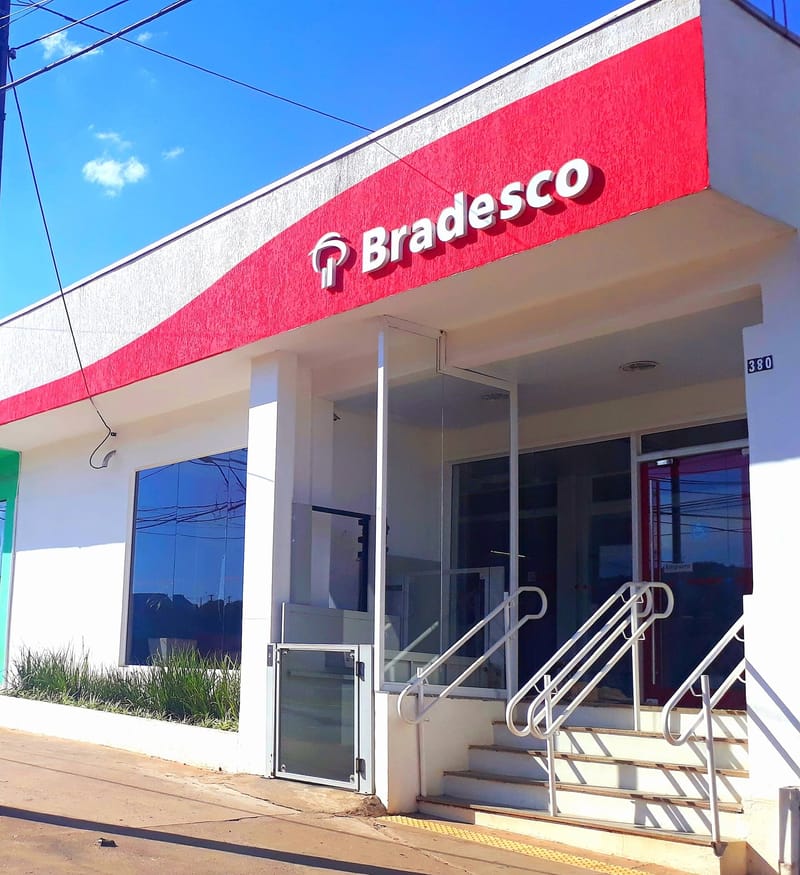 Fachada branca e vermelha de uma agência do Bradesco, com o nome do banco em letras brancas e escada até a porta de vidro