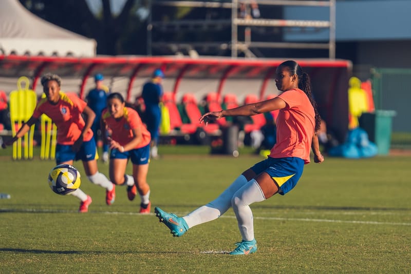 Jogadora da seleção brasileira feminina de base chuta a bola em treino, com colegas de camisa laranja ao fundo, em gramado sob luz de fim de tarde