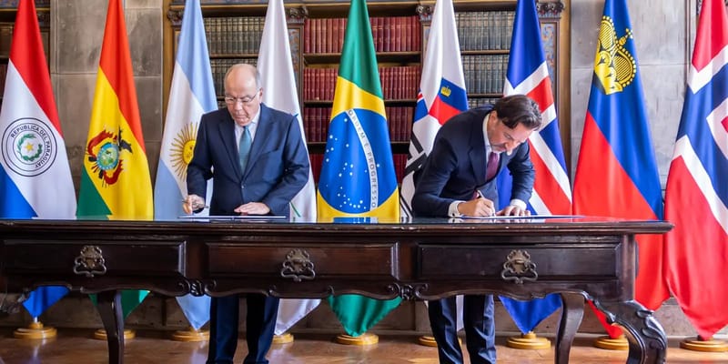 Dois homens de terno assinam documentos numa mesa de madeira, diante de bandeiras de Brasil, Noruega e outros países