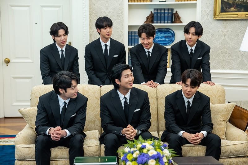 Os sete integrantes do BTS, de terno preto e gravata, quatro em pé atrás de um sofá bege e três sentados nele