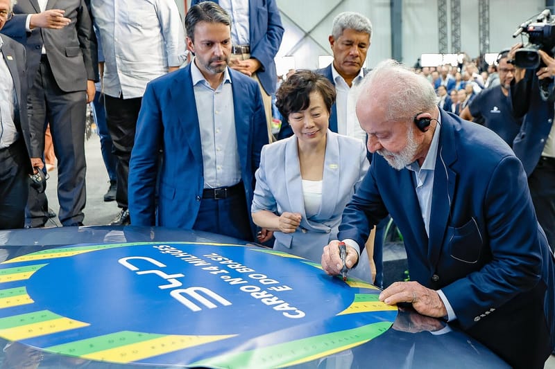 O presidente Lula, de terno azul, assina com caneta a superfície de um carro com o logotipo da BYD, cercado por autoridades e equipe de imprensa
