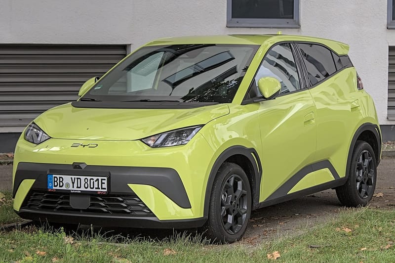 Hatch elétrico BYD verde-limão, visto de frente e de lado, estacionado sobre a grama diante de um prédio