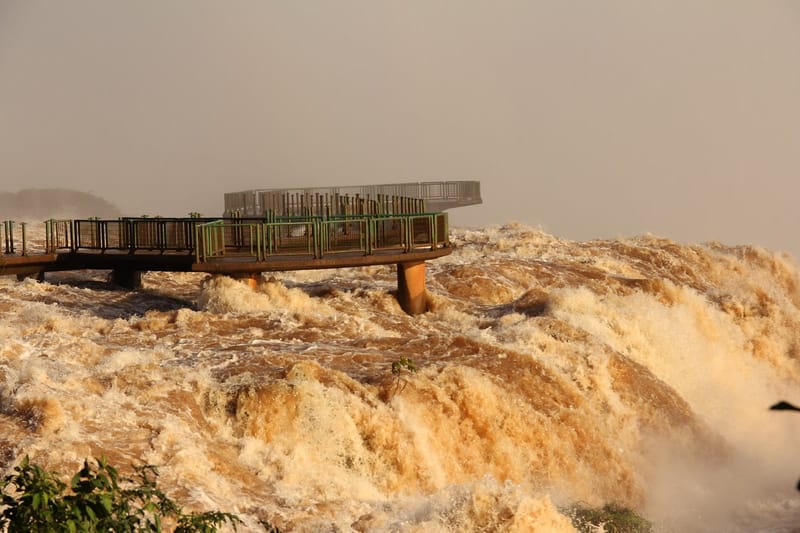 Passarela de metal com grades verdes avança sobre a água barrenta e espumante das Cataratas do Iguaçu em cheia