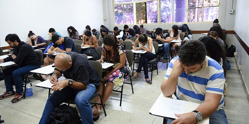 Candidatos sentados em carteiras escolares preenchem provas em uma sala de aula com janelas ao fundo